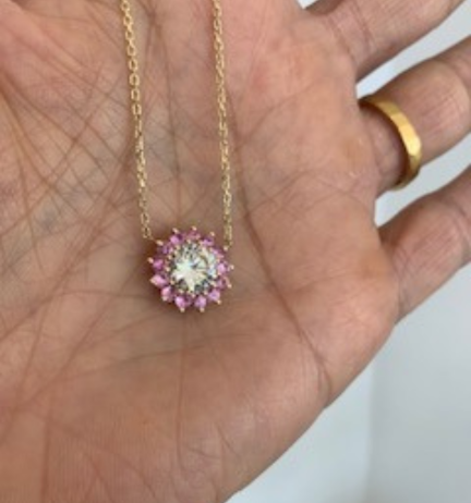Close-up of a hand holding a delicate gold necklace featuring a sparkling round pendant adorned with pink gemstones, highlighting its intricate design and elegance.