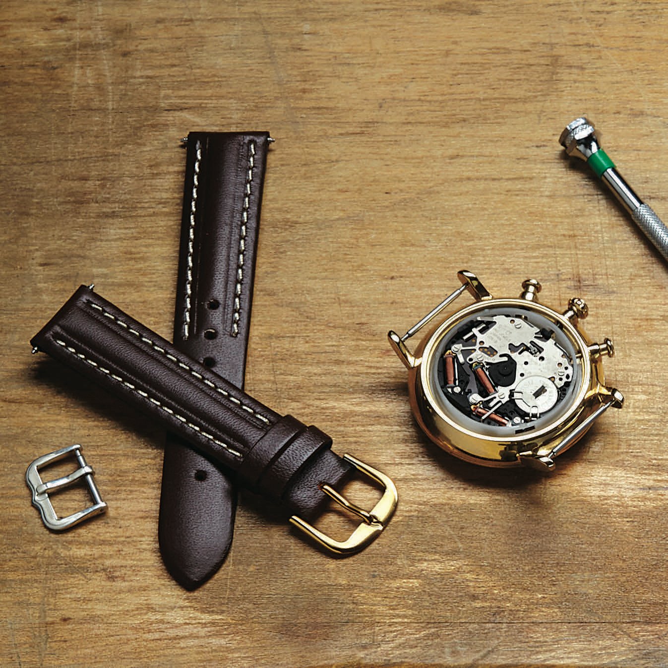 Close-up of a watch disassembled on a wooden surface, featuring a gold watch case, exposed mechanical movement, two dark brown leather watch straps with white stitching, and a silver buckle. A small screwdriver is also visible, indicating watch repair or assembly.