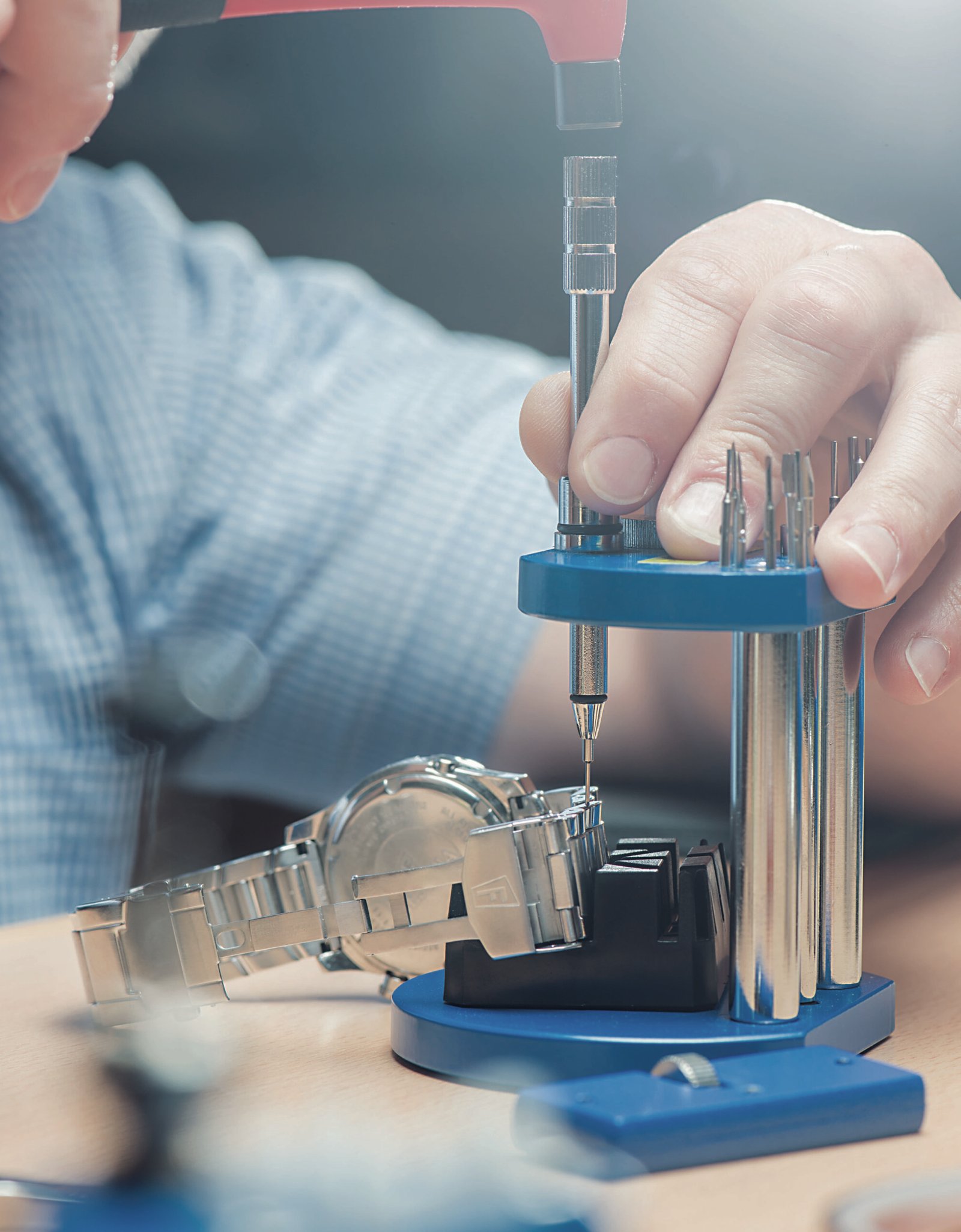 Close-up of a watch repair process, featuring a technician using a precision tool to adjust a watch strap on a blue workbench, surrounded by various watchmaking tools.