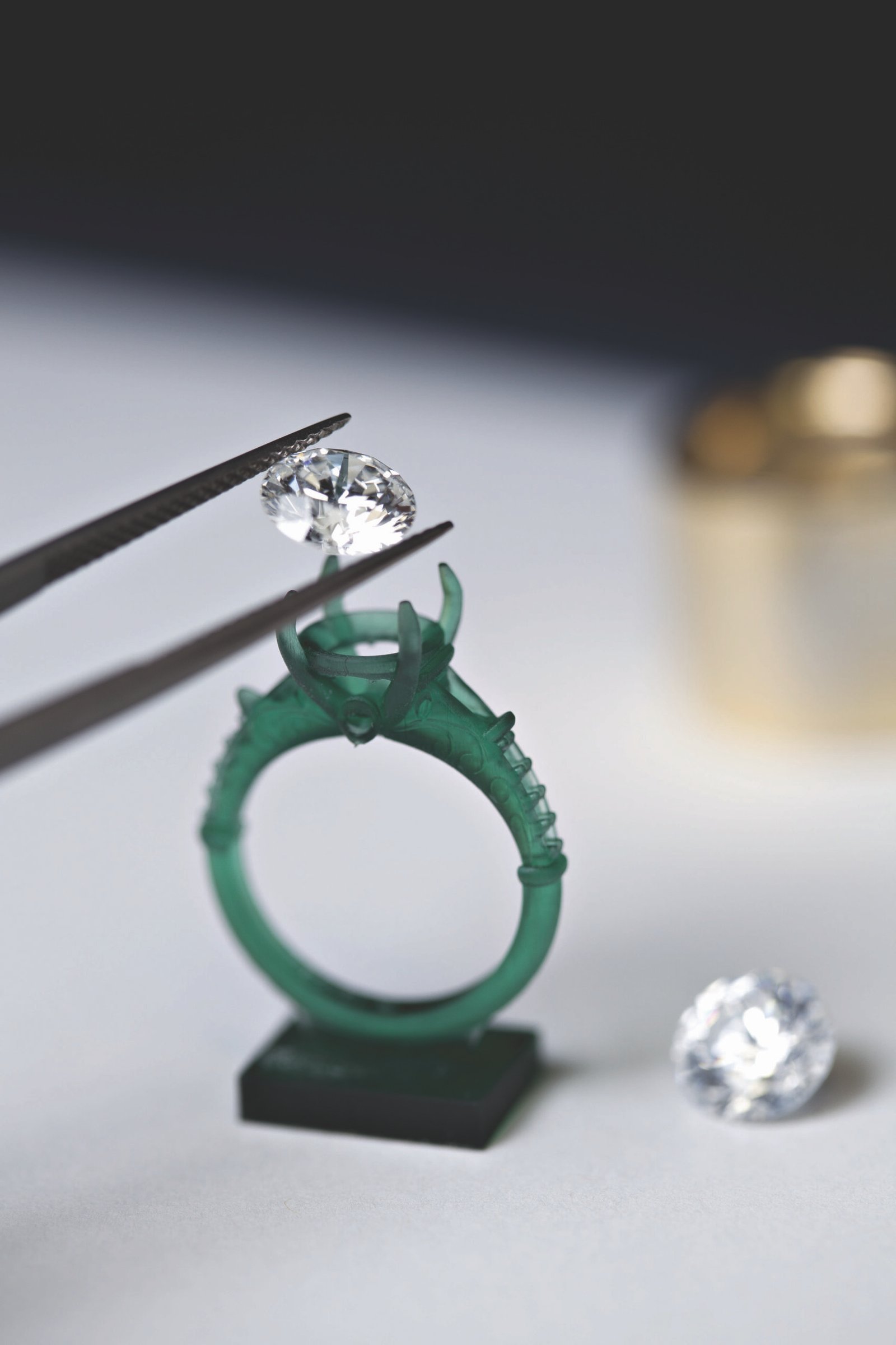 Close-up of a jeweler using tweezers to hold a sparkling diamond above a green wax ring model, with a second diamond resting nearby, illustrating the process of jewelry design and gemstone setting.