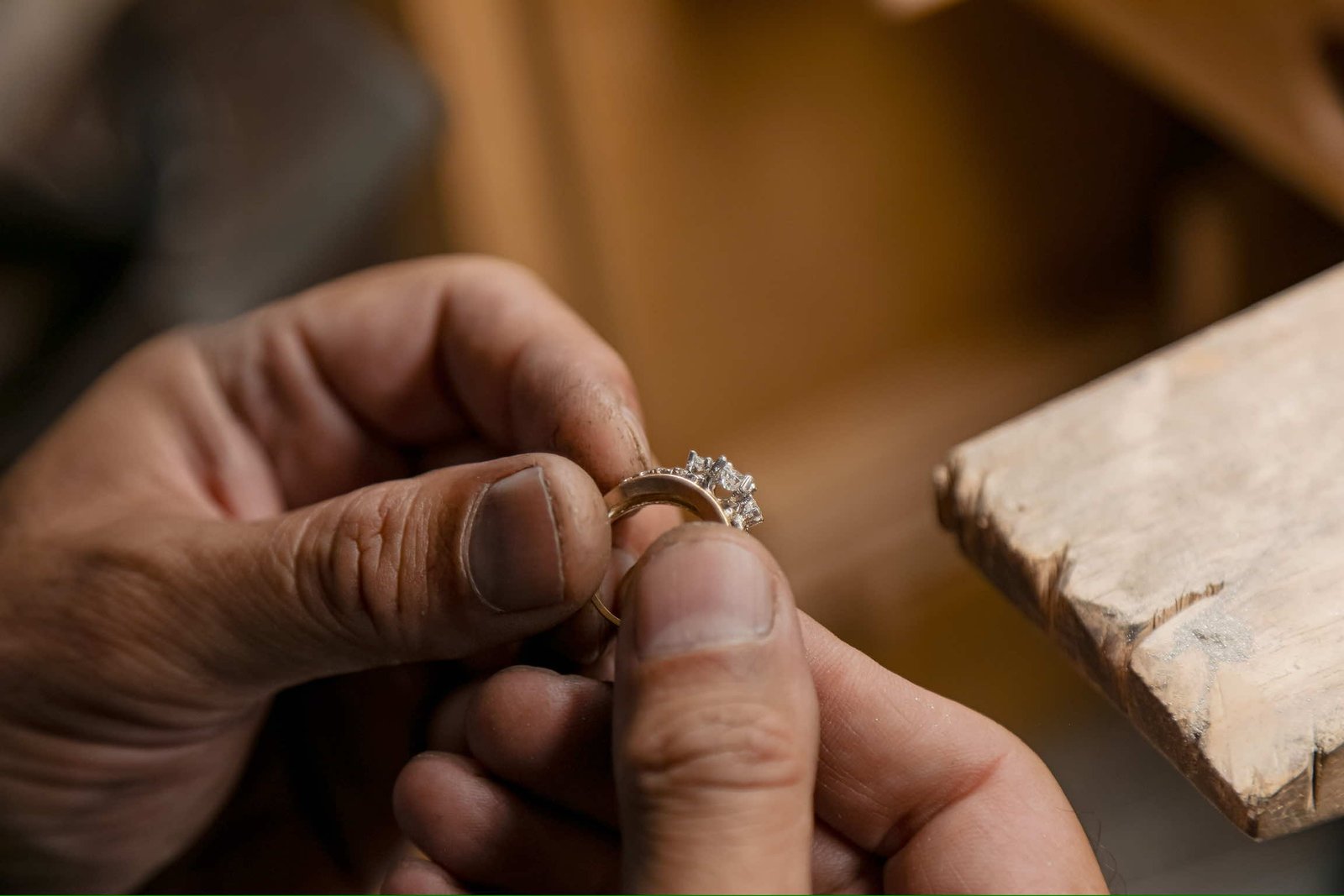 Close-up of a jeweler's hands holding a gold engagement ring with multiple diamonds, showcasing craftsmanship and attention to detail in a workshop setting.