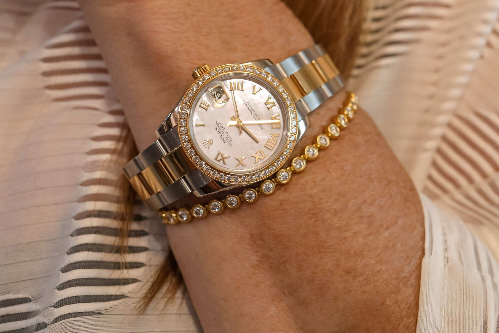 Close-up of a luxury two-tone Rolex watch with a diamond bezel and roman numerals, worn on a wrist adorned with a gold bracelet featuring sparkling stones. The background features soft, textured fabric.