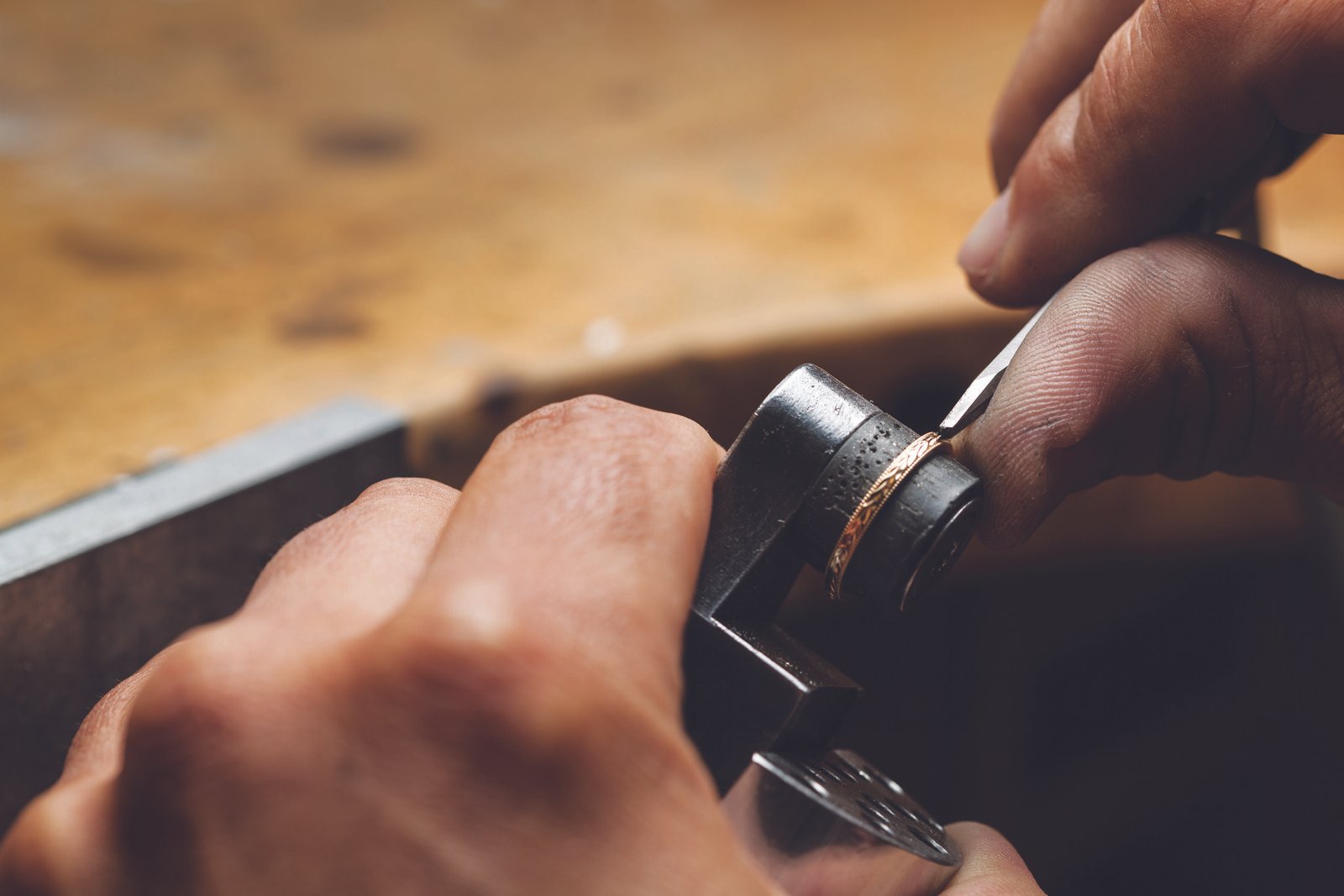 Close-up of a jeweler's hand using a tool to shape a delicate gold ring on a workbench, showcasing craftsmanship and attention to detail in jewelry making.
