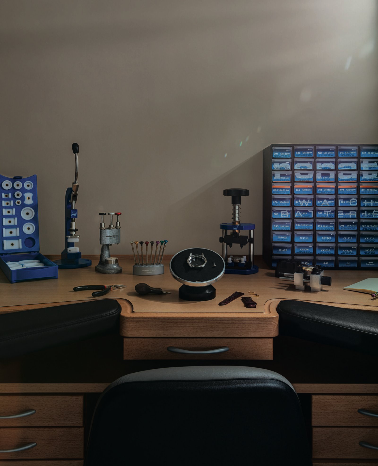 A well-organized watchmaking workspace featuring various tools and equipment, including a watch press, screwdrivers, and a display of watch parts, set on a wooden desk with black leather chairs.