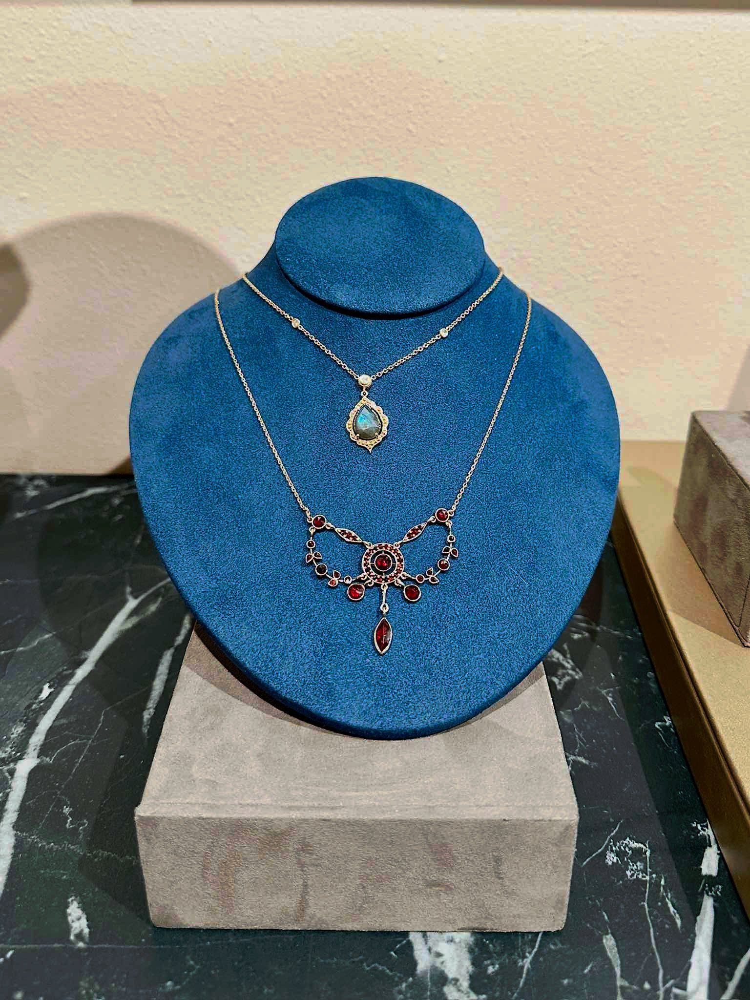 Elegant silver necklaces displayed on a blue velvet jewelry stand, featuring a teardrop pendant and a decorative piece with red gemstones. Perfect for showcasing fine jewelry in a retail setting.
