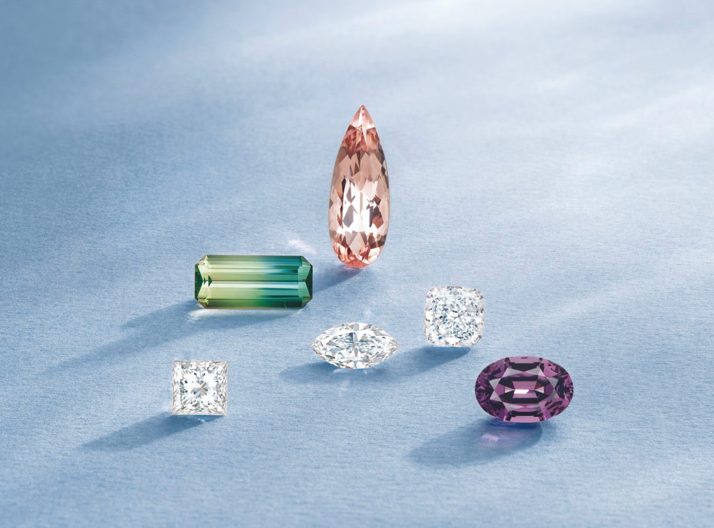 A collection of five colorful gemstones including a green tourmaline, a pink sapphire, a clear diamond, a purple spinel, and a white diamond, arranged artistically on a soft blue background.