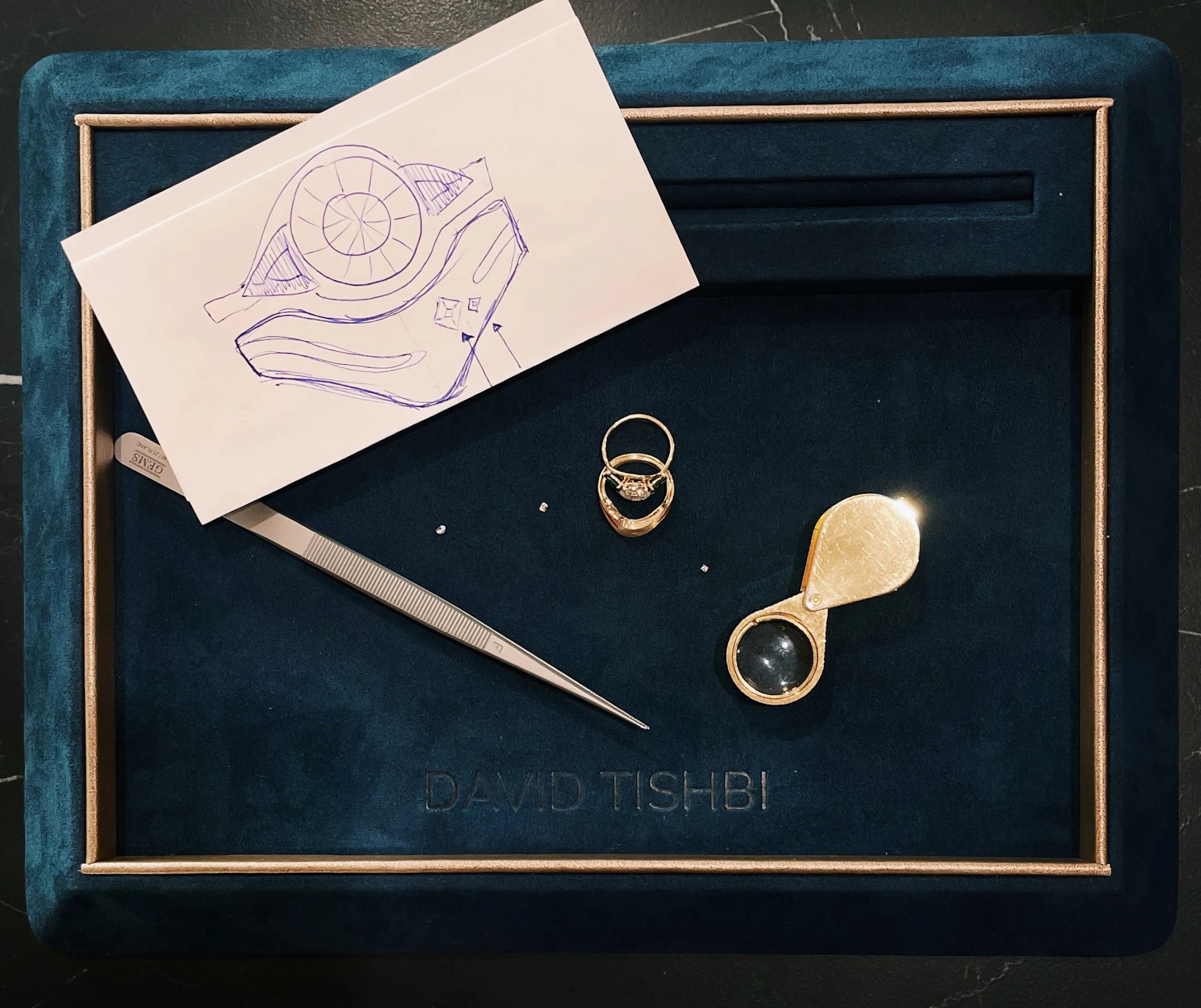 Jewelry design workspace featuring a sketch of a ring, a gold ring with a diamond, a magnifying loupe, and precision tweezers on a blue velvet tray embossed with "DAVID TISHBI." Ideal for showcasing custom jewelry creation and craftsmanship.